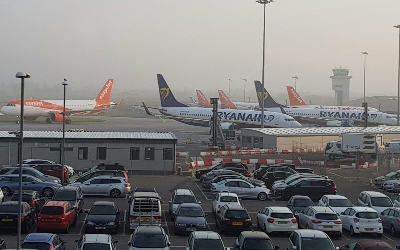 Photo of an airport carpark. Three stationary planes are in the background against a foggy sky with rows of parked cars in the foreground.