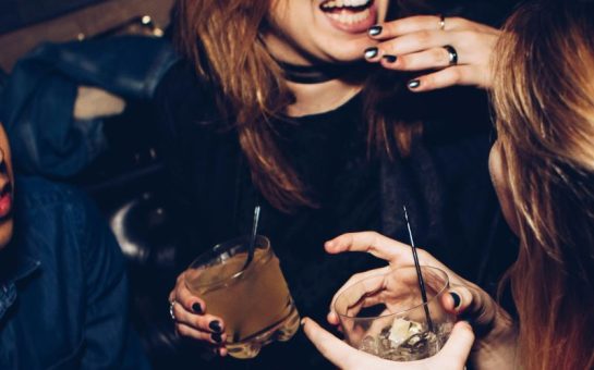 Two hands are shown holding glasses of alcohol. A woman's smile is shown, with the top of her face cut off, and her hand holding a drink next to her friends being the centre of the image.