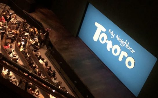 Image of a stage with a sky blue set background and white text reading 'My Neighbour Totoro'. A theatre audience is seated in front of the stage.