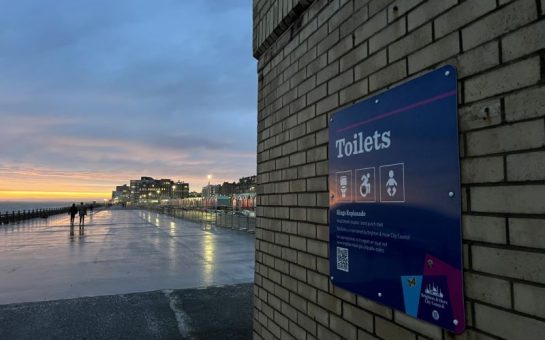 Landscape image of a boardwalk by Brighton's sea. On the right of the image is a bricked building with a sign titled 'toilets' on it. The left side of the image shows the length of the boardwalk, with the pebble beach running on the left edge of the image as a late sunset is shown in the background.