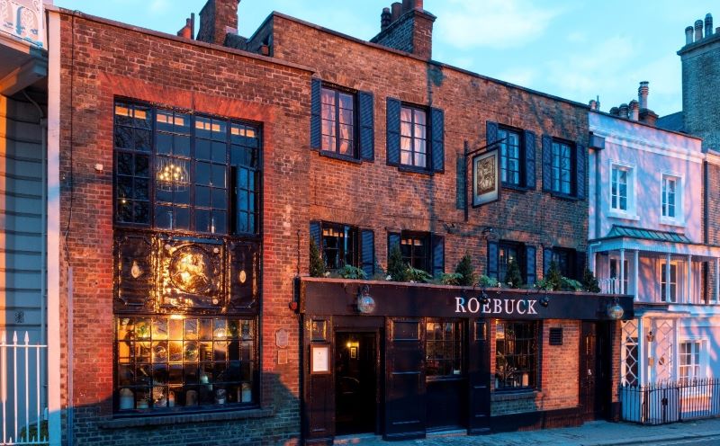 Image of a red brick pub with black window frames against a blue sky. The name of the pub 'Roebuck' hangs over the opening.