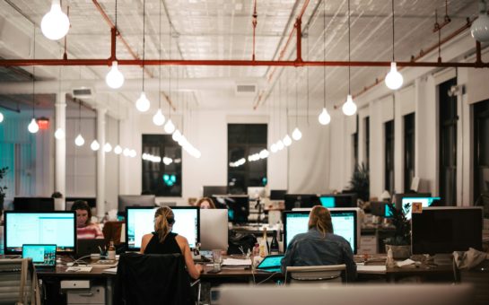 Image of an open plan office, High ceilings and tear drop shaped lights. People are sat on desks with their backs to the camera, working on computers.