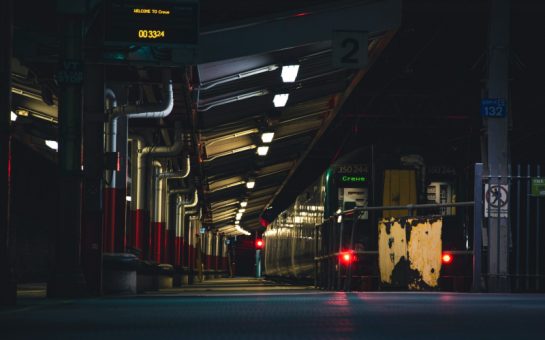 A photo in dark lighting shows a train station platform with a train with red lights pulling away from it. The glowy letters on the train sat Crewe and the platform is empty.