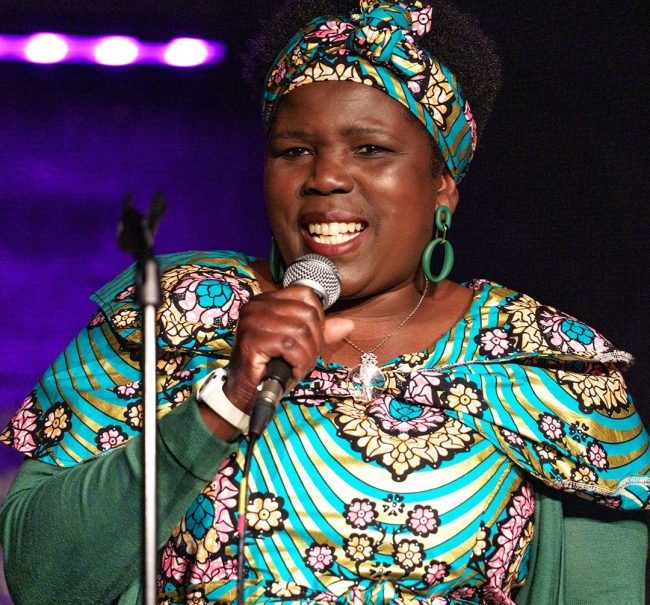 A woman stands in floral dress, holding a microphone, on a stage with lighting behind her.