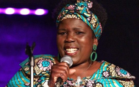 A woman stands in floral dress, holding a microphone, on a stage with lighting behind her.