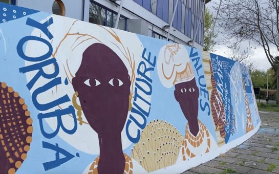 Mural in painted on a curved wall in blue, yellow, white, and purple. A building is visible behind and some pavement in front.