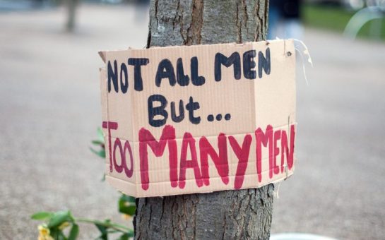 A cardboard sign painted saying not all men but too many men hung around a tree
