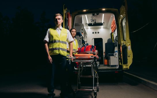 Two night paramedic shift workers stood infront of an ambulance at night.