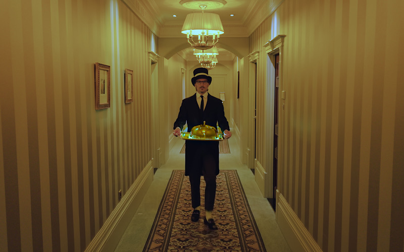 Oliver Proudlock dressed as a hotel bellboy walking down a corridor.