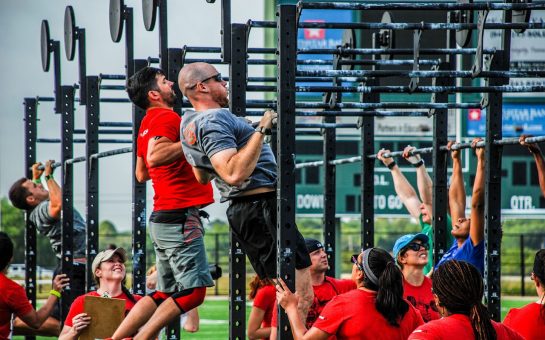 An image of six male CrossFit athletes completing a chest-to-bar pull up on an outside rig. Around them, there are judges watching their reps.