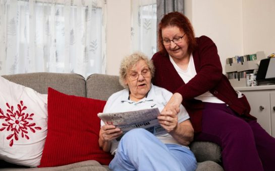 Photo of an elderly lady and a carer on a couch.
