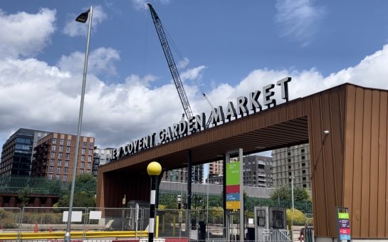 The New Covent Garden Market entrance runs next to the train tracks in Nine Elms, alongside a blue sky with clouds.