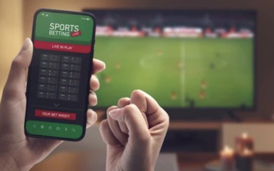 A person on a betting app with a football match on the TV in the background