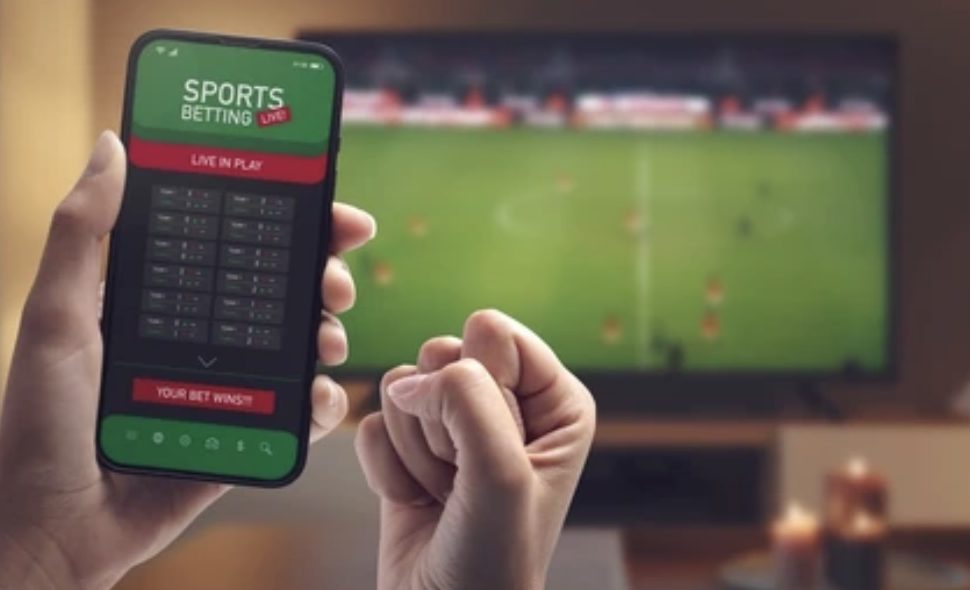 A person on a betting app with a football match on the TV in the background