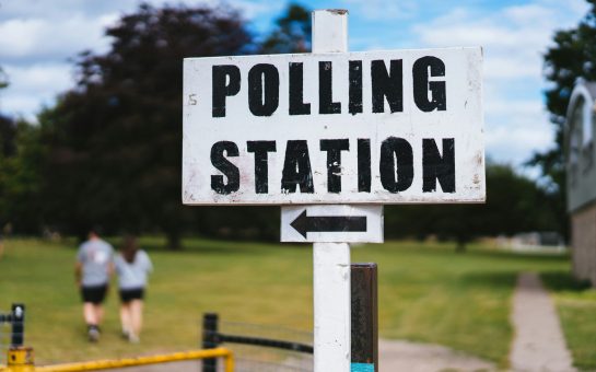 A white sign with black writing reads Polling Station with an arrow facing left with a park in the background.