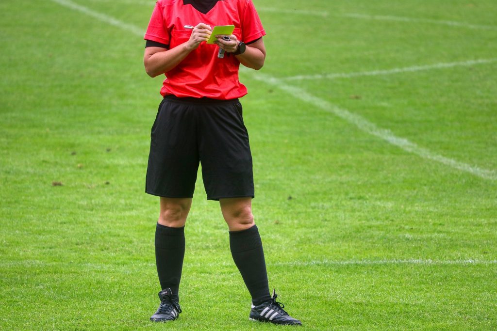 Football referee in black shorts and red shirt writing down a players' number on a yellow card after making a booking