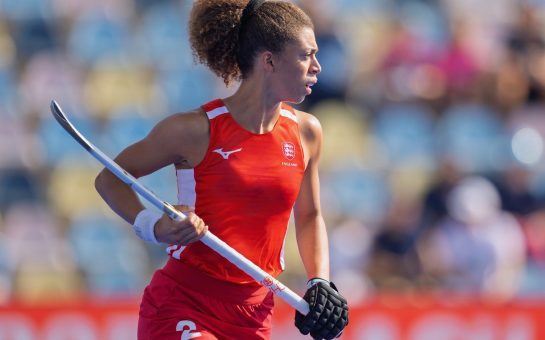 Darcy Bourne playing hockey for England. She wears a red vest and skort, and holds her stick in the air with two hands (Credit: Graeme Wilcockson (@gmwsportphoto on Instagram))