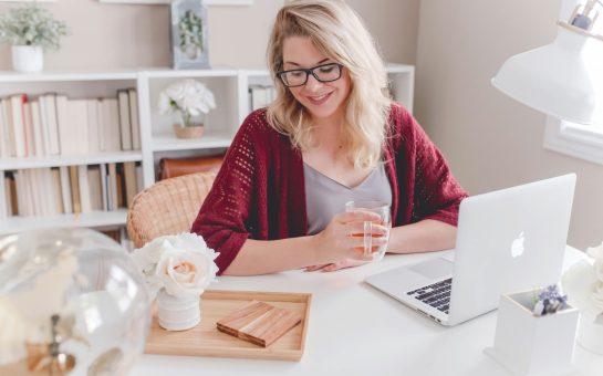 Woman smiling holding glass mug sitting beside table with MacBook (Credit: Free to use from Unsplash)