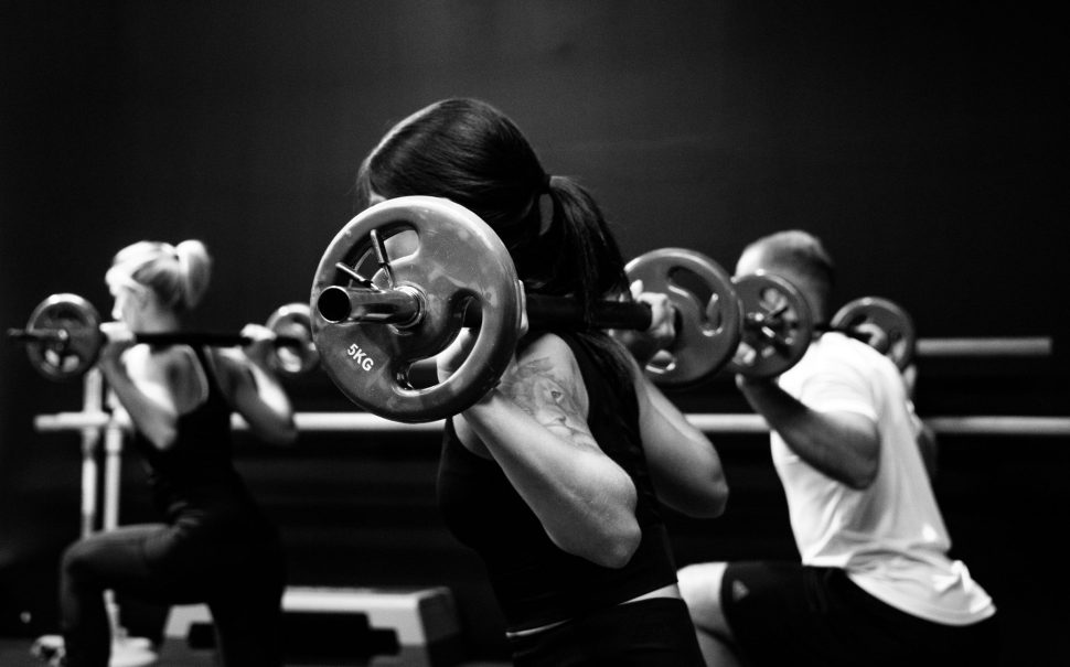 Black and white image of two women and one man in a gym with barbell weights on their shoulders performing a lunge