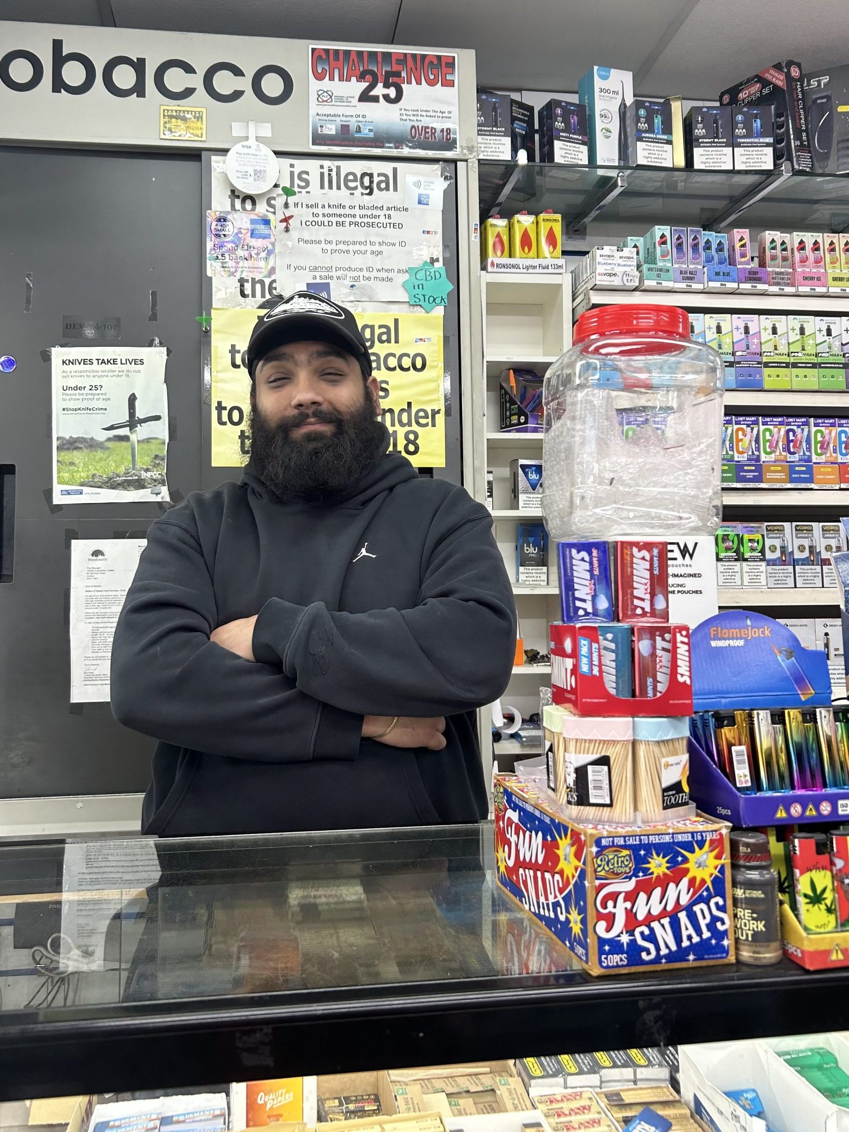Picture of a man called Anu with his arms crossed in a black hoodie taken in the local Tooting off license where he works.