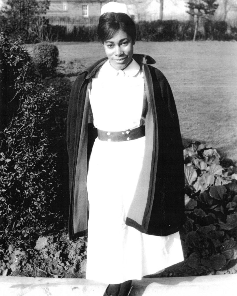 A black and white photograph of Windrush nurse Dawn hill. She wears a long nurses gown and a black cape.