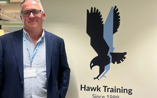 Crawford Knott dressed in a light blue shirt and dark blue blazer stood next to the Hawk Training logo - a hawk bird in flight