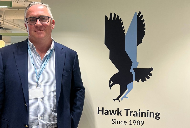 Crawford Knott dressed in a light blue shirt and dark blue blazer stood next to the Hawk Training logo - a hawk bird in flight