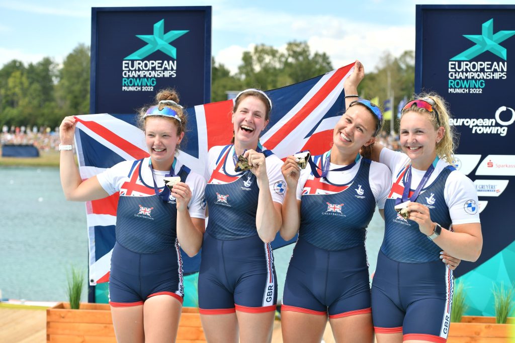 GB rowing four hold gold medal and GB flag on lakeside.