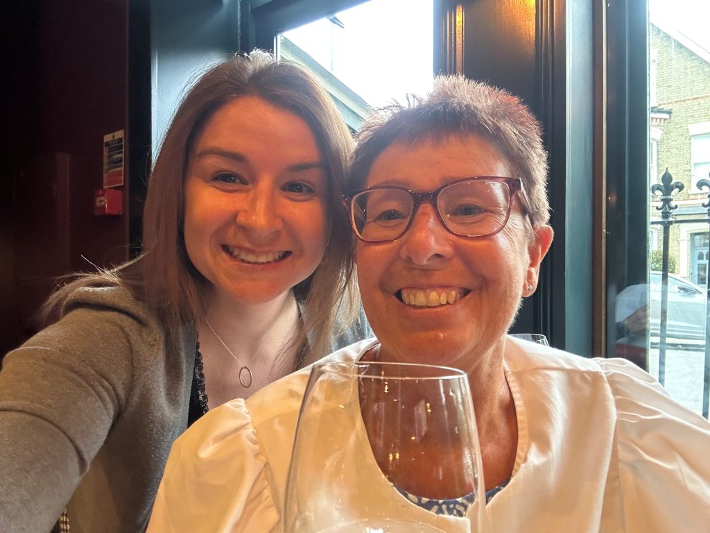 Me, Lucy, and my mum, Carol, smiling for a selfie at The Althorp on Bellevue Road. I have shoulder length brown hair, am wearing a black top and a grey cardigan. Mum has short brown/red hair, is wearing red glasses and is wearing a blue dress and a white cotton light jacket. You can see mum is holding a wine glass. We look cute.