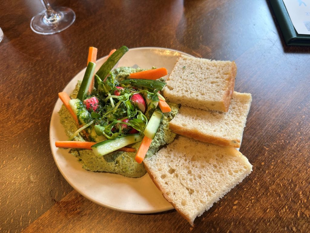 A plate of pea and mint humous with cucumber, carrots and radishes on top, and a side of focaccia. It looks fresh and crunchy.