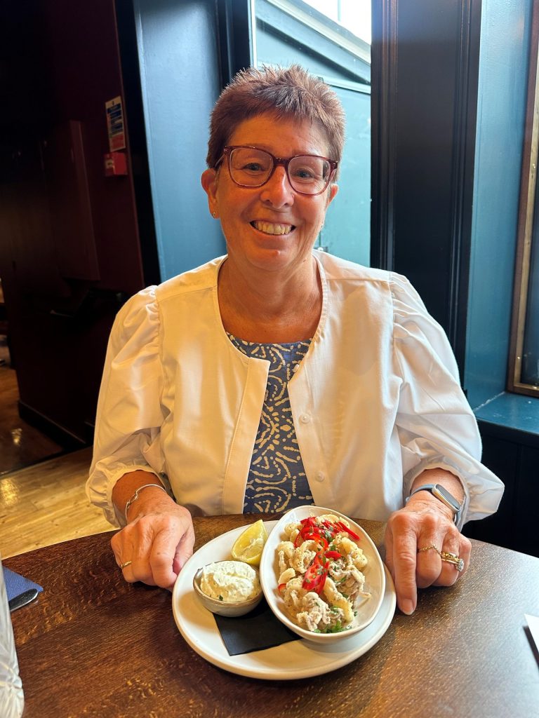 My mum Carol posing with her bowl of scampi for starter at the dinner table at The Althorp. Mum has short brown/red hair and red glasses. She is smiling for the camera. You can see she is wearing a blue dress and a white cotton light jacket.