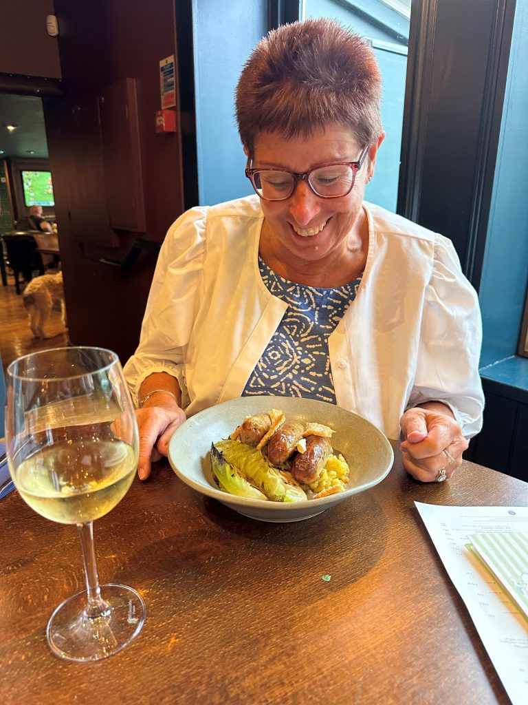 A picture of my mum Carol looking at her bowl of pork, leek & apple sausages, with crushed new potatoes, hispi cabbage, pork crackling and gravy at The Althorp. Mum has short brown/red hair and red glasses. You can see she is wearing a blue dress and a white cotton light jacket.