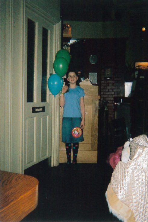 An 11-year-old Lucy at Dexter's Grill wearing a blue t-shirt, a denim skirt and spotty tights. I am standing and smiling for the camera holding three balloons.