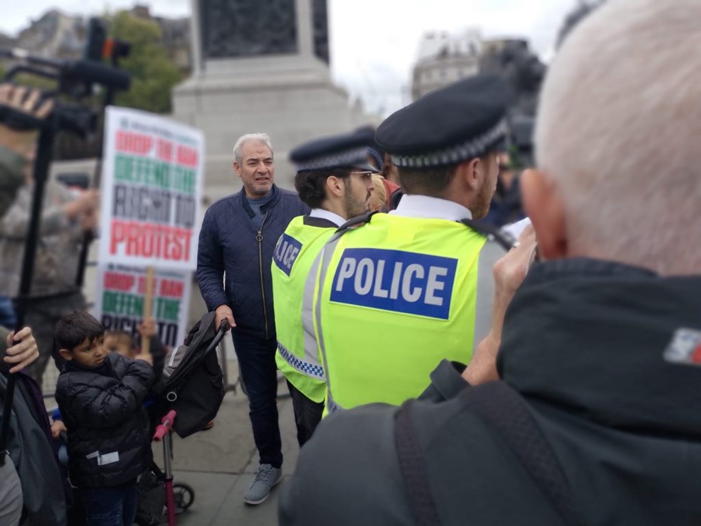 Police monitor the crowd in Trafalgar Square. A man with a pram, and a child holding a placard defending the right to protest, stand behind them. 