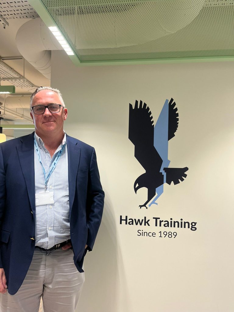 Crawford Knot dressed in a light blue shirt and dark blue blazer stood next to the Hawk Training logo - a hawk bird in flight
