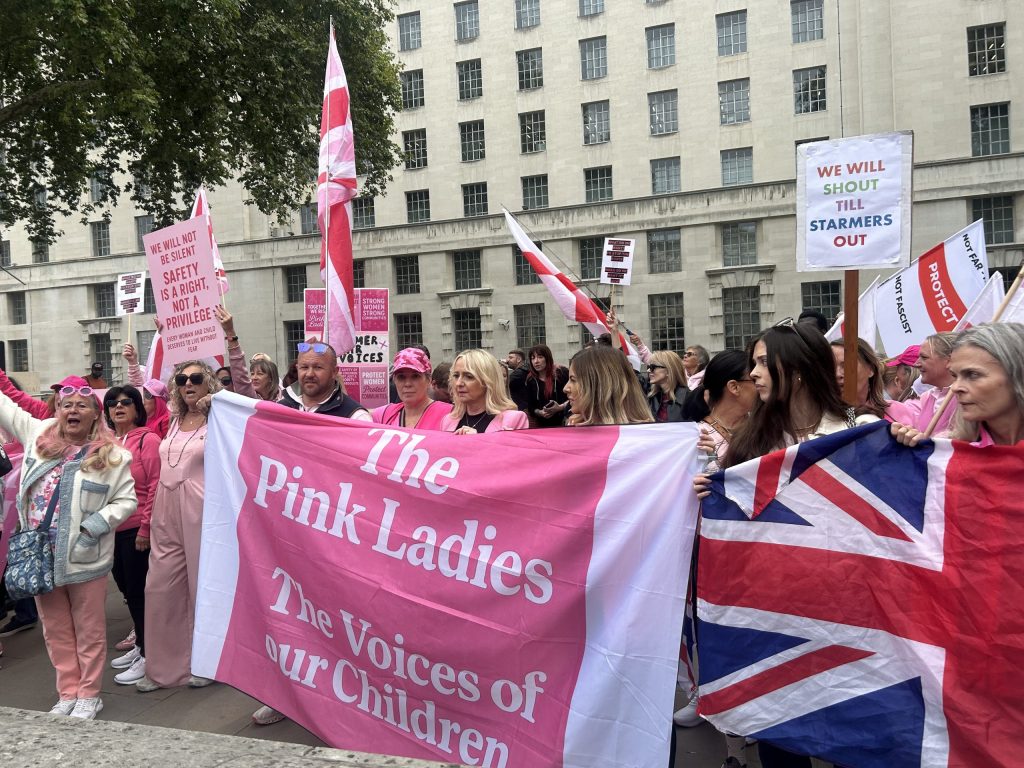 'Pink Lady' protestors hold sign that reads 'The Pink Ladies / The Voices of Our Children' in Westminster. On the right, a woman holds a Union Jack flag.
