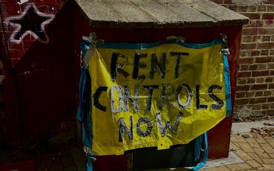 Blue and yellow sign on a table that reads 'Rent Controls Now' in bold silver writing, outside a meeting of the Newham branch of London Renters' Union (LRU).