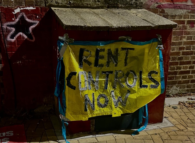 Blue and yellow sign on a table that reads 'Rent Controls Now' in bold silver writing, outside a meeting of the Newham branch of London Renters' Union (LRU).