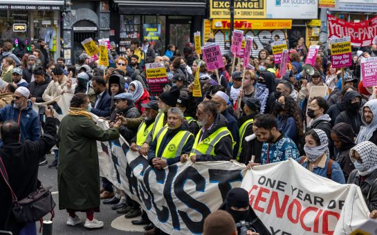 Crowds gather, some wearing yellow hi-vis, in a protest behind an obscured banner that reads "East London is Anti-Fascist" and another that says "one year of genocide". Other protesters hold pink signs saying "Refugees Welcome / Stop the Far Right".