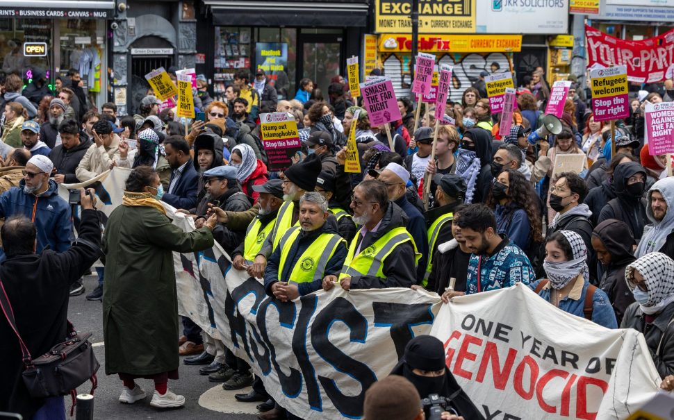 Crowds gather, some wearing yellow hi-vis, in a protest behind an obscured banner that reads "East London is Anti-Fascist" and another that says "one year of genocide". Other protesters hold pink signs saying "Refugees Welcome / Stop the Far Right".