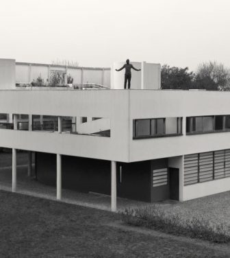 Black and white image of a man stood on top of a house, arms stretched out. The building is surrounded by trees.