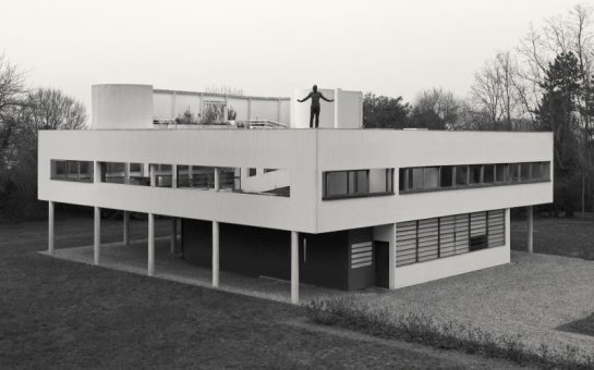 Black and white image of a man stood on top of a house, arms stretched out. The building is surrounded by trees.