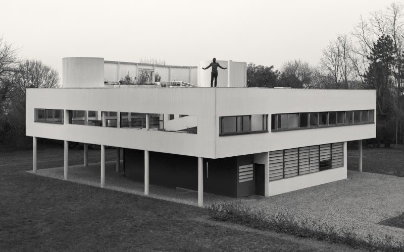 Black and white image of a man stood on top of a house, arms stretched out. The building is surrounded by trees.