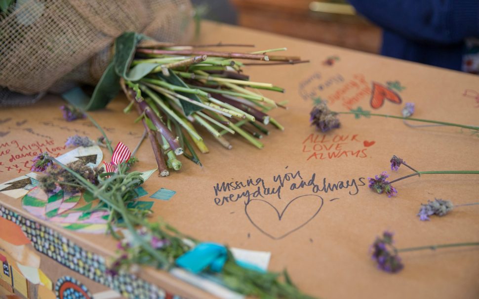 Cardboard coffin with handwritten tributes, flowers, and messages.