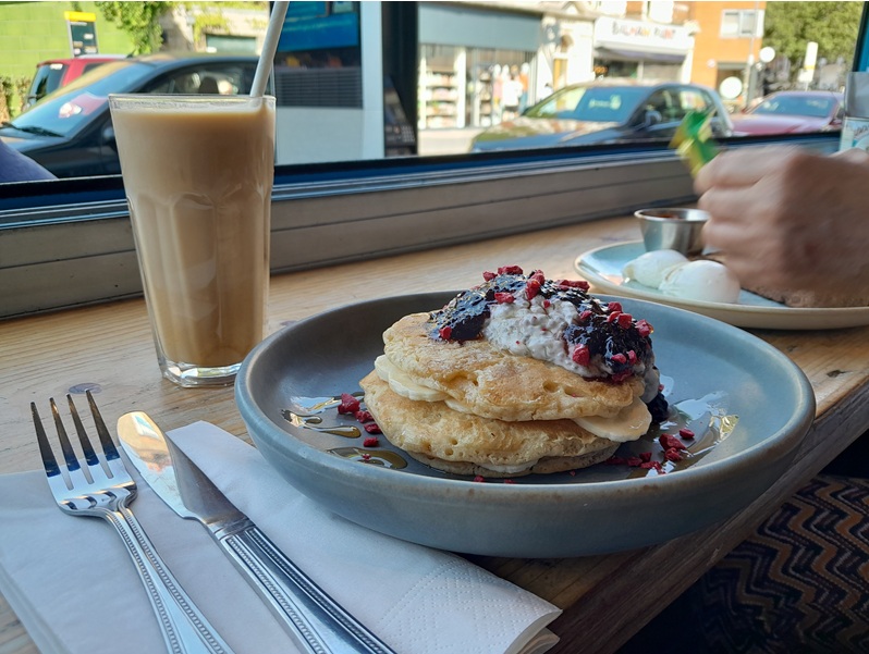 A stack of pancakes with berries and yoghurt on a plate next to a knife and folk and n iced coffee in the background.