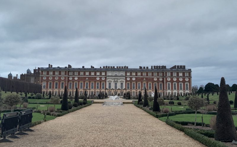 A grand, towering red brick palace with a long pebbled drive running up to a fountain, boarded by trees on either side.