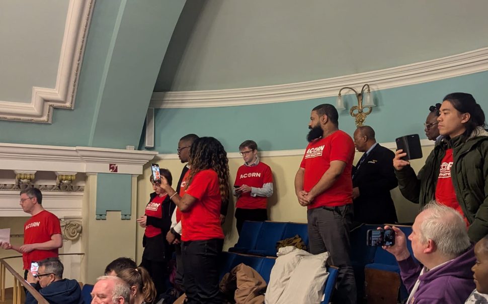 Members of ACORN Lambeth, stood in red ACORN Union t-shirts, stand in Lambeth Town Hall's public gallery.