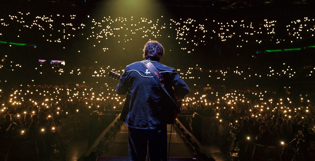 man on stage in front of a large crowd shining phone lights