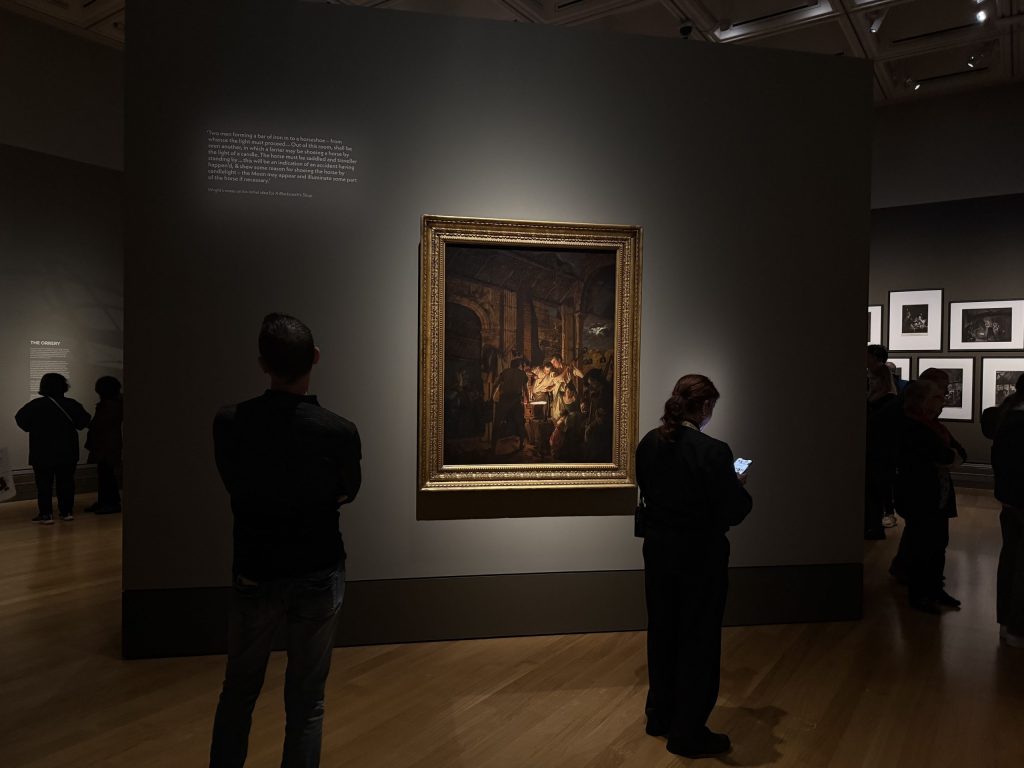 Visitors at the exhibition, one looks at a painting whilst the other looks down to her phone