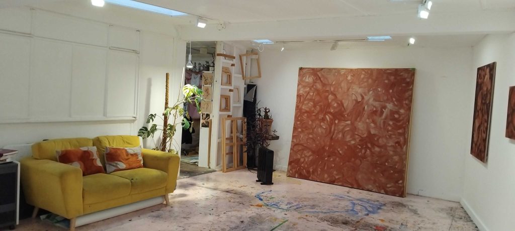 Photograph of art gallery showing a white room with a yellow sofa, large red piece of art work, and a floor covered in multi-covered paint splatters.